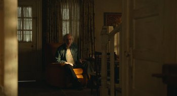 Movie still from “The Tender Bar” (2021), directed by George Clooney – An older man sitting on a chair in a room; Wide shot, Low angle