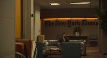 Movie still from “The Tender Bar” (2021), directed by George Clooney – A man sitting on a couch in front of the new york times logo; Wide shot, Over the shoulder angle