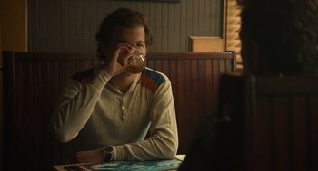 Movie still from “The Tender Bar” (2021), directed by George Clooney – A man sitting at a table drinking from a glass; Medium shot, Over the shoulder angle