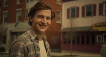 Movie still from “The Tender Bar” (2021), directed by George Clooney – A young man smiling for the camera in front of a brick building; Close Up shot, Over the shoulder angle