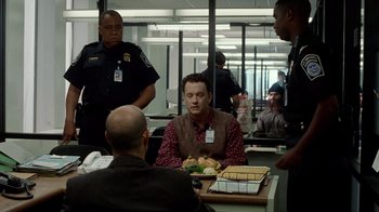 Movie still from “The Terminal” (2004), directed by Steven Spielberg – A group of men standing around a table with a plate of food on top of it; Medium shot, Over the shoulder angle