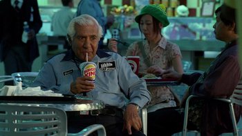 Movie still from “The Terminal” (2004), directed by Steven Spielberg – An older man sitting at a table with a drink; Medium shot, High angle