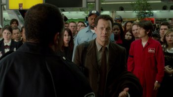 Movie still from “The Terminal” (2004), directed by Steven Spielberg – A man in a brown suit and tie standing in front of a group of people; Medium shot, Over the shoulder angle