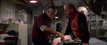 Movie still from “The Thing” (1982), directed by John Carpenter – Two men are sitting at a table and one is cutting a cake; Medium shot, Low angle