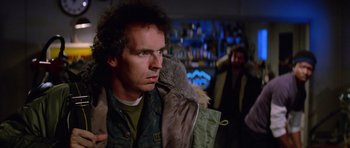 Movie still from “The Thing” (1982), directed by John Carpenter – A man holding a backpack in a room with other people; Close Up shot, Over the shoulder angle