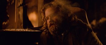 Movie still from “The Thing” (1982), directed by John Carpenter – A man with long hair and a beard is sitting in front of a fireplace; Medium shot, Low angle