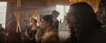 Movie still from “The Three Musketeers - Part I: D'Artagnan” (2023), directed by Martin Bourboulon – A group of people sitting in front of candles; Medium shot, Over the shoulder angle