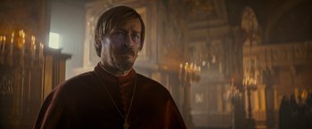 Movie still from “The Three Musketeers - Part I: D'Artagnan” (2023), directed by Martin Bourboulon – A man with a beard wearing a red robe and a cross necklace; Close Up shot, Over the shoulder angle