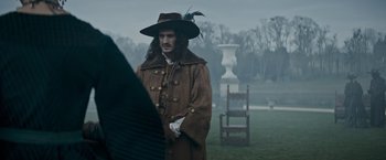 Movie still from “The Three Musketeers - Part I: D'Artagnan” (2023), directed by Martin Bourboulon – A man in a hat standing in the grass; Medium shot, Over the shoulder angle
