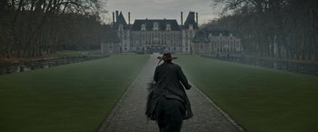 Movie still from “The Three Musketeers - Part I: D'Artagnan” (2023), directed by Martin Bourboulon – A man riding a horse down a path in front of a large building; Wide shot, Low angle