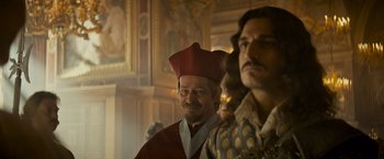 Movie still from “The Three Musketeers - Part I: D'Artagnan” (2023), directed by Martin Bourboulon – A man in a red hat standing next to another man in a red hat; Medium shot, Over the shoulder angle