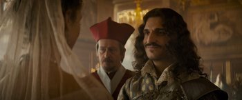 Movie still from “The Three Musketeers - Part I: D'Artagnan” (2023), directed by Martin Bourboulon – A man with long hair is standing next to a priest; Close Up shot, Over the shoulder angle