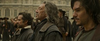 Movie still from “The Three Musketeers - Part I: D'Artagnan” (2023), directed by Martin Bourboulon – An old man with long hair is standing in front of a group of people; Close Up shot, Over the shoulder angle