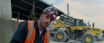Movie still from “The Town” (2010), directed by Ben Affleck – A man in an orange vest and a hard hat in front of a large wheel loader; Medium shot, Low angle
