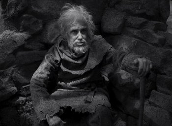 Movie still from “The Tragedy of Macbeth” (2021), directed by Joel Coen – An old man sitting on a pile of rocks with a cane; Medium shot, High angle