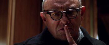 Movie still from “Enemy of the State” (1998), directed by Tony Scott – An older man wearing glasses and holding his index finger to his mouth; Close Up shot, Low angle