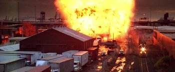 Movie still from “Enemy of the State” (1998), directed by Tony Scott – A large fire is burning in the background of a warehouse; Extreme Wide shot, High angle