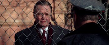 Movie still from “Enemy of the State” (1998), directed by Tony Scott – An older man wearing a suit and tie standing in front of a chain link fence; Close Up shot, Over the shoulder angle
