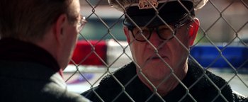 Movie still from “Enemy of the State” (1998), directed by Tony Scott – A man wearing a police hat is looking at another man through a chain link fence; Close Up shot, Over the shoulder angle