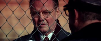 Movie still from “Enemy of the State” (1998), directed by Tony Scott – An older man wearing a suit and tie behind a chain link fence; Close Up shot, Over the shoulder angle