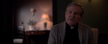 Movie still from “The Unholy” (2021), directed by Evan Spiliotopoulos – An older man wearing a clerical collar and standing in front of a lamp; Close Up shot, Over the shoulder angle