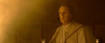 Movie still from “The Unholy” (2021), directed by Evan Spiliotopoulos – A man wearing a priest's outfit in a dimly - lit room; Close Up shot, Low angle