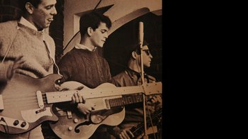 Movie still from “The Velvet Underground” (2021), directed by Todd Haynes – A man holding a guitar in front of a microphone; Medium shot, Overhead angle