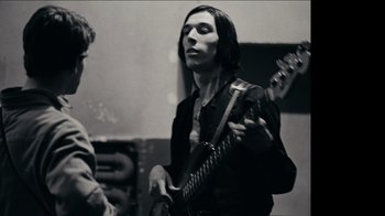 Movie still from “The Velvet Underground” (2021), directed by Todd Haynes – A man holding a guitar while standing next to another man; Medium shot, Low angle