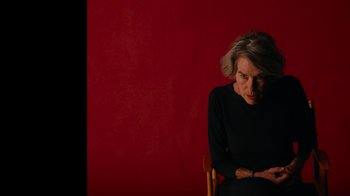 Movie still from “The Velvet Underground” (2021), directed by Todd Haynes – An older woman sitting in a chair in front of a red wall; Close Up shot, Over the shoulder angle