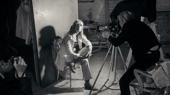 Movie still from “The Velvet Underground” (2021), directed by Todd Haynes – A woman sitting on a chair in a room with a camera; Medium shot, High angle