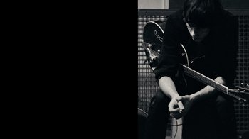 Movie still from “The Velvet Underground” (2021), directed by Todd Haynes – A man playing a guitar while sitting on a chair; Medium shot, High angle