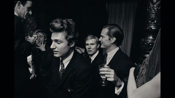 Movie still from “The Velvet Underground” (2021), directed by Todd Haynes – A group of men standing next to each other holding drinks; Medium shot, Over the shoulder angle