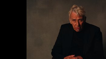 Movie still from “The Velvet Underground” (2021), directed by Todd Haynes – An older man sitting in a chair in front of a gray wall; Close Up shot, Low angle