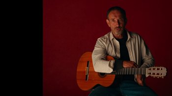 Movie still from “The Velvet Underground” (2021), directed by Todd Haynes – An older man sitting in front of an acoustic guitar; Medium shot, Low angle