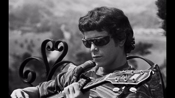 Movie still from “The Velvet Underground” (2021), directed by Todd Haynes – A man holding a microphone and a guitar in his hand; Close Up shot, Low angle