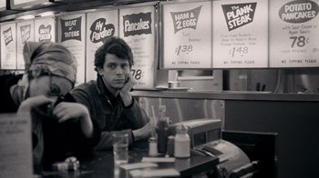 Movie still from “The Velvet Underground” (2021), directed by Todd Haynes – A black and white photo of a man sitting at a table in a restaurant; Medium shot, Over the shoulder angle
