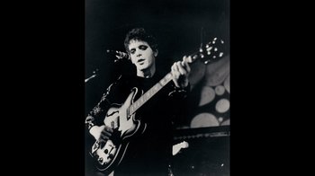 Movie still from “The Velvet Underground” (2021), directed by Todd Haynes – A man holding a guitar while standing on a stage; Medium shot, Low angle