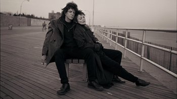 Movie still from “The Velvet Underground” (2021), directed by Todd Haynes – A man and a woman sitting on top of a wooden bench; Medium shot, Low angle