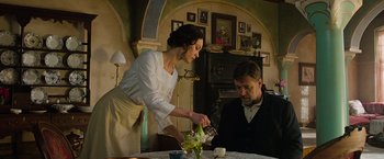 A woman in a white apron and a man in a black suit are at a dining table