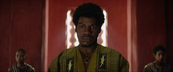 Movie still from “The Woman King” (2022), directed by Gina Prince-Bythewood – A man in a yellow and black shirt is looking at the camera; Close Up shot, Low angle