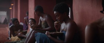 Movie still from “The Woman King” (2022), directed by Gina Prince-Bythewood – A group of people sitting on a bench eating food; Medium shot, Over the shoulder angle