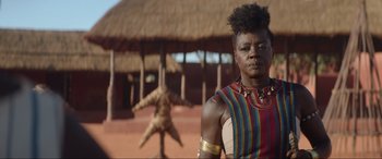 Movie still from “The Woman King” (2022), directed by Gina Prince-Bythewood – A woman in a colorful shirt standing in front of a building; Close Up shot, Over the shoulder angle