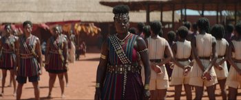 Movie still from “The Woman King” (2022), directed by Gina Prince-Bythewood – A woman in a colorful dress standing on a dirt field; Medium shot, Over the shoulder angle