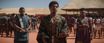 Movie still from “The Woman King” (2022), directed by Gina Prince-Bythewood – A man in a robe and a gold chain standing in front of a crowd; Medium shot, Over the shoulder angle