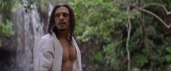 Movie still from “The Woman King” (2022), directed by Gina Prince-Bythewood – A man with long black hair is standing in front of trees; Medium shot, Over the shoulder angle