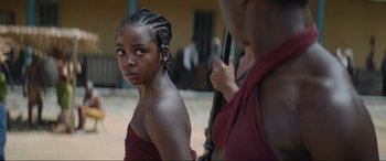 Movie still from “The Woman King” (2022), directed by Gina Prince-Bythewood – A young woman in a red dress is looking at the camera while a man in a red shirt is holding a gun; Close Up shot, Over the shoulder angle