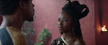 Movie still from “The Woman King” (2022), directed by Gina Prince-Bythewood – A woman with a black and gold dress and a black hat; Close Up shot, Over the shoulder angle