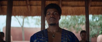 Movie still from “The Woman King” (2022), directed by Gina Prince-Bythewood – A man wearing a blue and gold shirt and a necklace; Close Up shot, Low angle