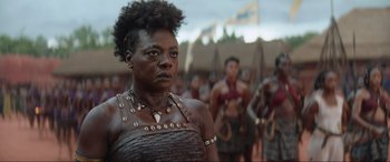 Movie still from “The Woman King” (2022), directed by Gina Prince-Bythewood – A woman in a costume stands in front of a group of people; Close Up shot, Over the shoulder angle