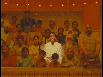 Movie still from “The Wonderful Story of Henry Sugar” (2023), directed by Wes Anderson – A group of people sitting in front of an image of a man; Wide shot, High angle